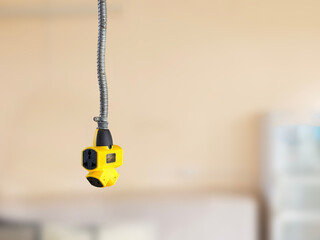A yellow multi-outlet plug with black sockets hangs from a flexible conduit against a blurred beige background.