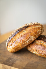 Two loaves of sourdough bread, freshly baked and still warm, are stacked on a wooden surface. The loaves are covered in seeds sesame and sunflower, adding a rustic and flavorful touch.