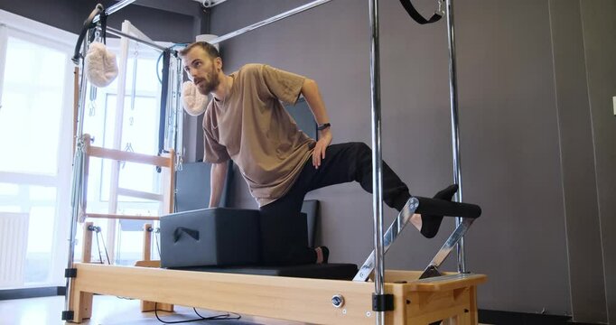 A young light-skinned man reshaping his body through exercises on a Reformer Pilates machine in a Pilates studio