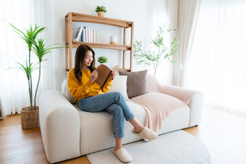 Young beautiful Asian woman reading a book on couch at home