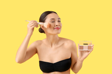 Fototapeta premium Young asian woman applying face powder with makeup brush and contour palette on yellow background, her eyes are closed in concentration, and she wears a strapless top