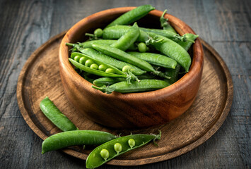 General plan of green young peas in pods in a wooden bowl on a wooden tray, on a gray textured wooden table. Low key.