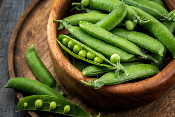 Young green peas in pods in a wooden bowl on a wooden tray on a gray wooden table. Side and top view, close-up.
