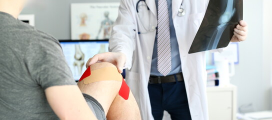 Close-up of smart physician in white uniform and striped tie checking damaged ankle. Troubled joint bound with protective red patches to prevent blood infection. Medical treatment concept