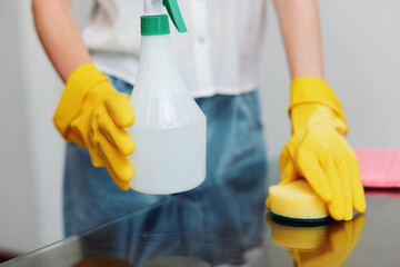 Woman in yellow rubber gloves holding cleaning solution and sponge on glass table in front of...