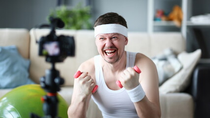 Male vlogger holds dumbbell in hand look and shout at camera on his subscribers, motivating them to action. Tutoring grade on an individual program online frauds concept aganist home background.