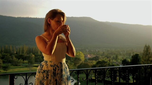 Woman stands on balcony with mountain view, enjoying cup of tea while appreciating tranquil morning light. Scenic serene moment. Relaxation and peaceful lifestyle in nature