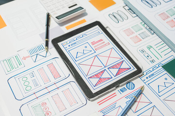 On a desk with paper, a tablet, and a laptop, a digital designer works on user interface (UI) and...