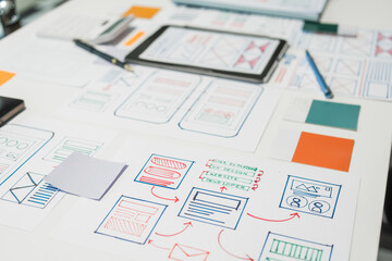 On a desk with paper, a tablet, and a laptop, a digital designer works on user interface (UI) and...