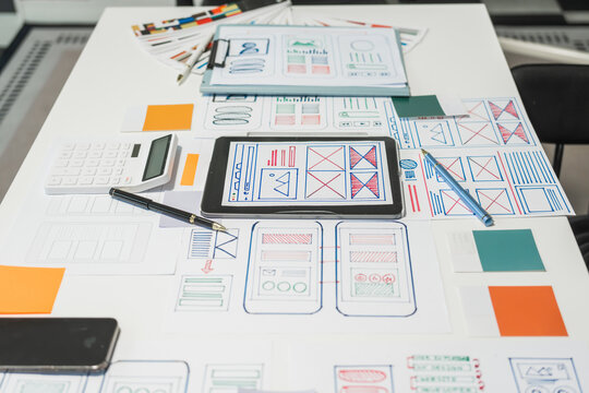 On a desk with paper, a tablet, and a laptop, a digital designer works on user interface (UI) and user experience (UX) concepts, focusing on usability, accessibility, and design principles.