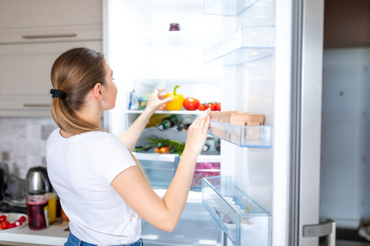 Portrait of female standing near open fridge full of healthy food, vegetables and fruits.