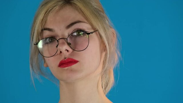 Female model smiles at device emitting positive energy. Young woman wearing glasses squints making suspicious look on blue background slow motion