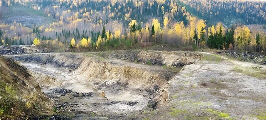Wide shot of a large open pit mining site in the Canadian woods during autumn. Trees can be seen on the horizon and dirt is visible at the bottom of mine shafts