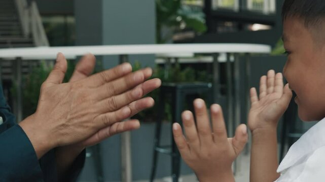 Father and small son dressed in school uniform clap hands in synchronized rhythm. Man and charming schoolboy sit at table in cozy cafe and play lively hand-clapping game