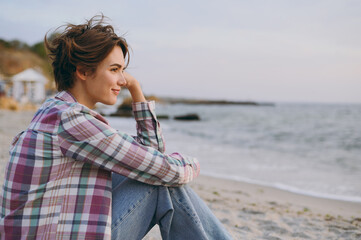 Side view young calm minded pensive thoughtful woman wear shirt casual clothes sits on plaid look aside rest on sea ocean sand shore beach outdoor seaside in summer day free time. Lifestyle concept.