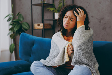 Little sick ill sad kid teen girl wrapped in plaid hold head suffer from headache sits on blue sofa couch cough stay home hotel flat spend free spare time in living room. Health, cold season concept