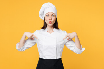 Young surprised chef cook baker woman she wear white shirt black apron uniform toque chefs hat point index finger down on area mock up isolated on plain yellow background studio. Cooking food concept.