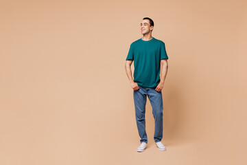 Full body young smiling fun happy cheerful middle eastern man wear blue t-shirt casual clothes look aside on area mock up isolated on plain pastel beige background studio portrait. Lifestyle concept.