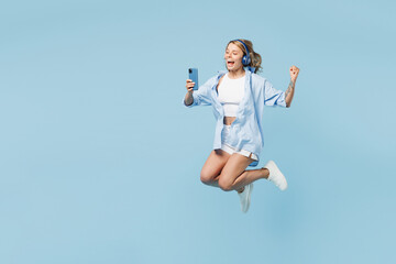 Full body young woman wears white top shirt casual clothes jump high listen to music in headphones use mobile cell phone do winner gesture isolated on plain pastel blue background. Lifestyle concept.