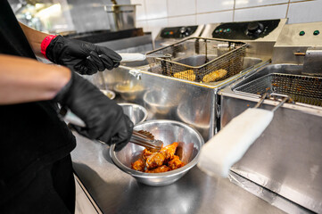 A person’s hands, wearing black gloves, use tongs to place fried chicken wings into a metal bowl in a commercial kitchen