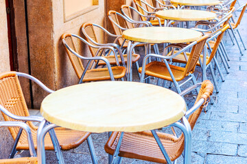 Row of empty tables with chairs on the street. The chairs are made of wood and metal. The tables are round and have a white top. The chairs are arranged in a row, with some of them facing the street