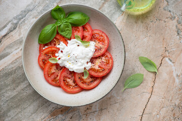 Plate with sliced tomatoes, stracciatella cheese and fresh basil, above view on a beige and roseate granite background, horizontal shot
