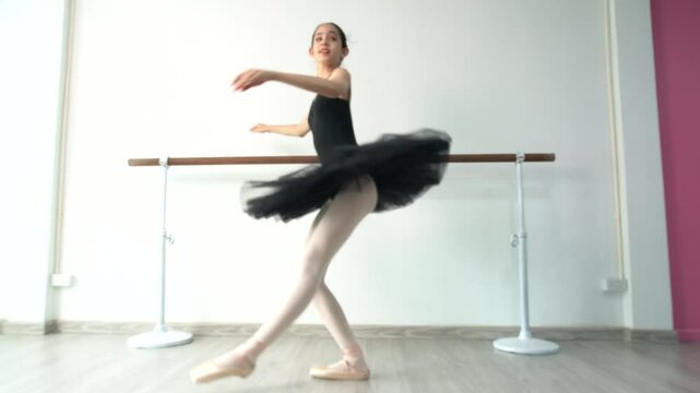 Young beautiful fourteen year old girl in black ballet tutu practicing ballet dancing practicing dancing as a black swan using bar pole, white wall in studio