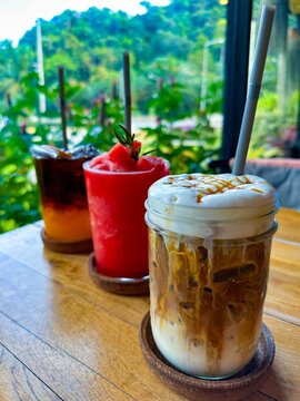 A selection of refreshing drinks including iced coffee, watermelon smoothie, and citrus iced tea on a wooden table, set against a lush tropical backdrop. Perfect for food and beverage themes.