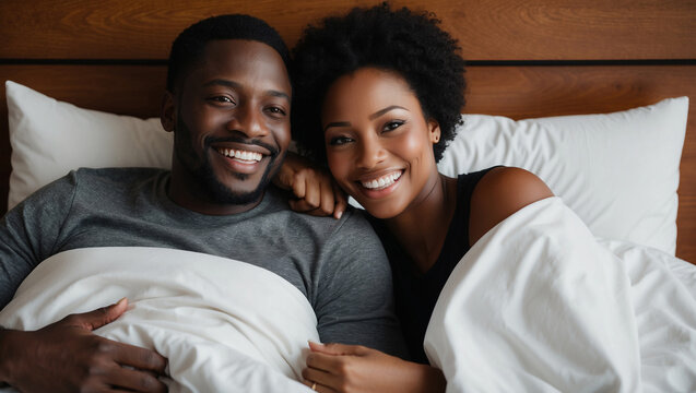 Happy black couple lies in bed together, smiling and looking at camera. They are both covered by white sheets and pillows