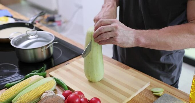 Man cuts zucchini in half on wooden board. Vegetables such as corn tomatoes onions and cucumber placed nearby on table foreshadow vegetarian dish slow motion
