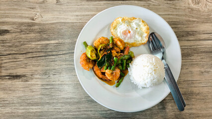 Stir-fried shrimp with chili paste over steamed rice on a white plate on a wooden table. Thai food.