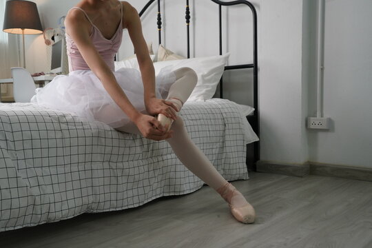 Young beautiful fourteen year old teenage girl sitting on bed in white ballet tutu practicing ballet dancing and wearing tied ribbed shoes in bedroom at home