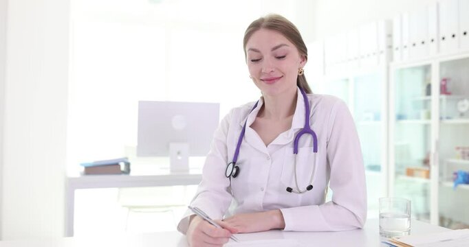 Woman with stethoscope around neck and in medical gown sits at table smiling. Female doctor in medical office waiting for patient slow motion