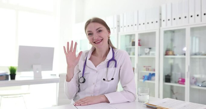Female doctor dressed in medical gown sits at table smiling. Cheerful woman at medical office waves hand waiting patient slow motion