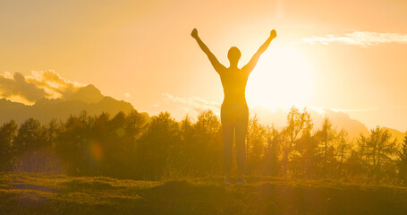 SILHOUETTE, LENS FLARE Athletic lady with raised arms on mountaintop at sunset