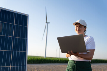 Woman farmer wearing white cap and t-shirt with laptop stands next to solar panel. Wind turbines in the background