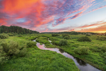 A winding river flows through a verdant meadow, bathed in the golden light of a beautiful sunset....