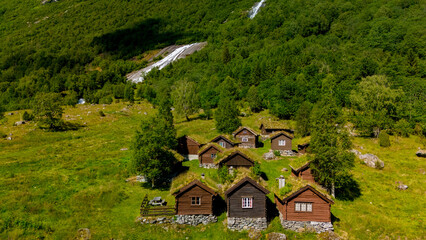 A cluster of charming wooden cabins with grass roofs dot a grassy hillside in Norway, with a cascading waterfall flowing down the forested mountainside behind them. lovatnet lake Lodal valley Norway