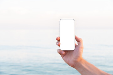 Close-up white screen Caucasian man holding a regular smartphone in his left hand, ocean beach in the background. High quality photo. Smartphone screen mockup cut out