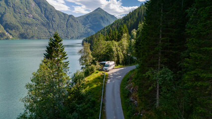 A white campervan parks on a winding road overlooking a picturesque Norwegian fjord, surrounded by lush green forests and mountains. Fjaerlandsfjorden, Fjord, Vestland, Norway