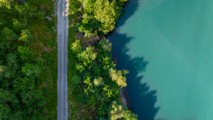 An aerial view of a winding road through lush green forest, leading to a turquoise lake in Norway. The sun casts long shadows across the water. Fjaerlandsfjorden, Fjord, Vestland, Norway