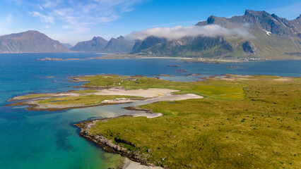 An aerial view of a small, green island with a sandy beach, surrounded by clear blue water and mountains in the distance. Kolbeinsanden Beach, Lofoten