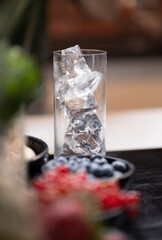 Cocktail glass filled with ice on black countertop. Berries and fruit in bowls on foreground. Cooling glass for cocktail or mocktail