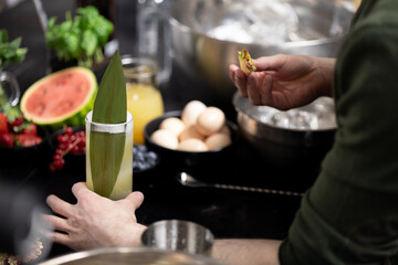 Barman preparing refreshing cocktail, adding green leaf garnish to tall glass. Drink surrounded by various ingredients, including watermelon, strawberries, eggs and ice