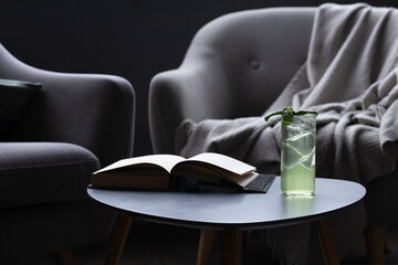 Glass of iced drink with sprig of mint on round table next to book. Gray armchair with throw...