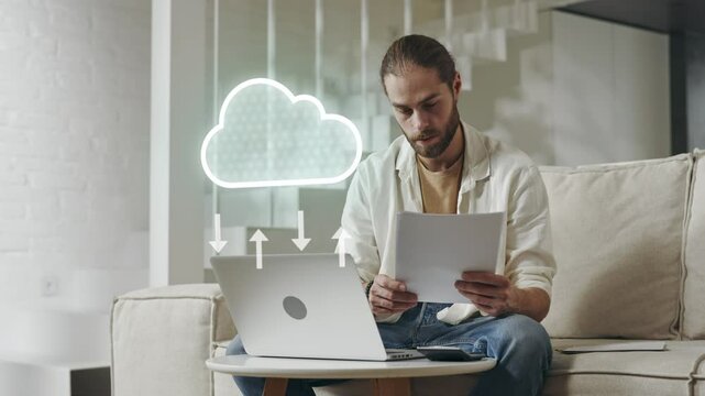 Modern Computer with Cloud Data Transfer Animation. Caucasian Man Working with Laptop and Documents at home Sitting on the Sofa.