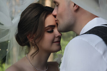 portrait of happy bride and groom under white veil, groom kisses the bride sensually and tenderly, wedding ceremony