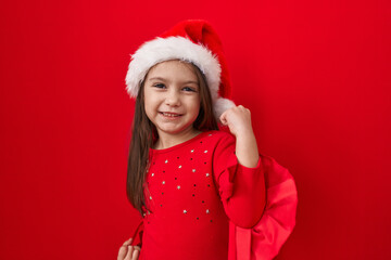 Joyful hispanic girl celebrating winning success, wearing adorable christmas hat, eyes closed and excited arms over head, isolated on bright red background