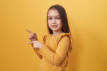 Adorable hispanic girl smiling, looking confidently at the camera, cheerfully pointing with two hands to side, over yellow isolated background