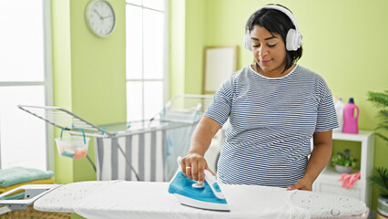 A hispanic woman enjoys music on headphones while ironing clothes in a colorful laundry room.